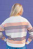 A woman from behind wearing a cream-colored knit sweater with horizontal stripes in orange and purple, standing against a purple painted brick wall.