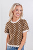 A woman wearing a brown short-sleeve knit top with white polka dots and cream ruffle trim at the collar, cuffs, and hem.