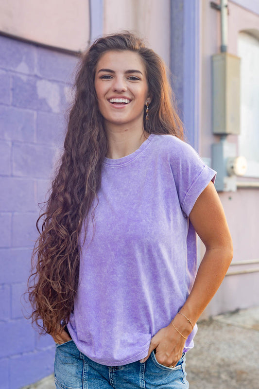 A smiling woman, wearing a short-sleeve crewneck t-shirt in a washed purple color, paired with blue jeans, standing against a purple brick wall.