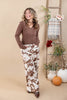 A woman modeling a brown v-neck top and brown-and-white cow-print pants in a warm autumn-themed studio setting with a ladder, plaid throw, pumpkins, and floral decor.