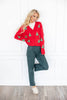 A woman models a festive red cardigan sweater featuring green Christmas tree patterns, paired with dark green pants and white loafers.