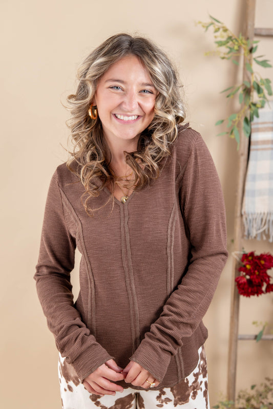 A smiling woman with wavy blonde hair wearing a brown waffle-knit V-neck long sleeve top and brown-and-white patterned pants, standing against a warm beige background with decorative plants.