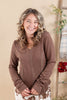 A smiling woman with wavy blonde hair wearing a brown waffle-knit V-neck long sleeve top and brown-and-white patterned pants, standing against a warm beige background with decorative plants.