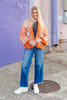 A woman stands outdoors on a sidewalk wearing an orange knit cardigan with white sequin ghost prints, blue jeans, a black top, and white sneakers, in front of a purple brick wall.