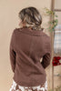 A woman viewed from behind wearing a brown ribbed knit long-sleeve top with a high collar and a central seam, paired with a white and brown patterned skirt, set against a warm beige background with decorative plants.