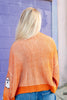 A woman viewed from behind wearing an orange and white vertically striped knit sweater with a white ghost patch on the sleeve, paired with blue jeans, standing against a purple brick wall.