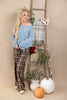 A woman stands in a styled studio setting wearing an off-the-shoulder light blue sweater and brown plaid pants. She is posed beside a wooden ladder with a plaid throw blanket, flowers, greenery, white vases, and pumpkins against a warm beige backdrop.