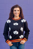 A woman wearing a black crewneck sweatshirt with white pumpkin faces and orange and black striped trim, standing against a purple brick wall.