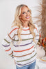A woman wearing a white knit sweater with horizontal stripes in orange, green, brown, and purple, paired with blue jeans.