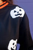 Close-up of a black crewneck sweatshirt featuring white textured jack-o'-lantern motifs on the chest and sleeve, worn by a woman with an orange striped collar visible.