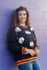 A woman wearing a black crewneck sweatshirt with white pumpkin faces and orange and black striped trim, leaning against a purple brick wall.