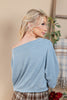 A woman shown from the back wearing a heathered light blue off-the-shoulder long-sleeve top, paired with brown and white plaid pants, in a warm beige indoor setting.