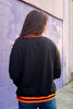 A woman viewed from behind wearing a black sweatshirt with bright orange ribbed trim at the collar and hem, paired with blue jeans, standing against a purple painted brick wall.