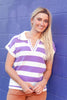 A smiling blonde woman wearing a purple and white horizontally striped short-sleeve polo shirt, standing against a purple wall.