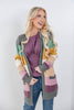 A smiling blonde woman models a long, open-front knit cardigan with large color-blocked panels in gray, sage green, mustard yellow, cream, pink, and mauve over a mauve top and blue jeans, against a plain light gray studio background.