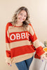 A woman wearing a ribbed knit sweater with bold horizontal stripes in rust orange and beige, featuring the word 'GOBBLE' in white lettering across the chest. She is also wearing blue jeans, standing against a solid beige background.