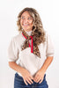 A woman with wavy blonde hair smiling and looking to the side, wearing a beige puff-sleeve top, blue jeans, and a leopard print neck scarf tied with a red ribbon, set against a plain white background.