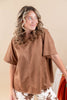 A woman wearing a brown faux suede short-sleeve top and white patterned pants, standing against a plain beige background.