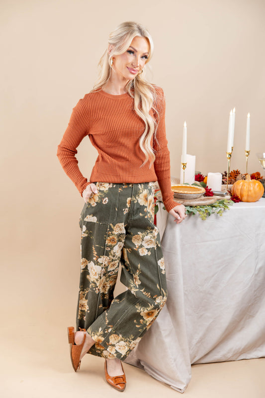A woman models a ribbed rust-orange long-sleeve top and wide-leg olive green pants with a beige floral pattern, standing next to a decorated table with pumpkins and candles.