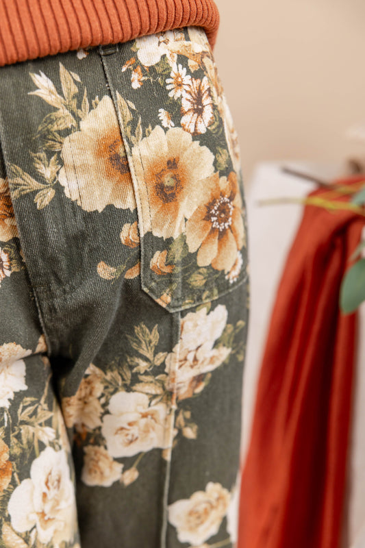 Close-up of a pair of dark olive green denim pants featuring a large cream and orange floral print, styled with an orange ribbed knit top.