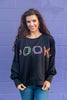A woman wearing a black crewneck sweatshirt with 'SPOOKY' spelled out in orange, green and purple plaid lettering, paired with blue jeans, standing against a purple brick wall.