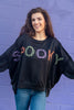 A woman wearing a black crewneck sweatshirt with 'SPOOKY' spelled out in orange, green and purple plaid lettering, paired with blue jeans, standing against a purple brick wall.