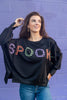A woman wearing a black crewneck sweatshirt with 'SPOOKY' spelled out in colorful plaid lettering, paired with blue jeans, standing against a purple brick wall.