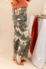 A woman wearing wide-leg floral pants in a muted sage green with cream and peach flowers, paired with an orange ribbed sweater and brown shoes, standing next to a draped table.