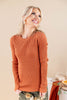 A smiling blonde woman wearing a ribbed long-sleeve crewneck sweater in a warm rust orange color, paired with floral pants, posed against a solid beige background.