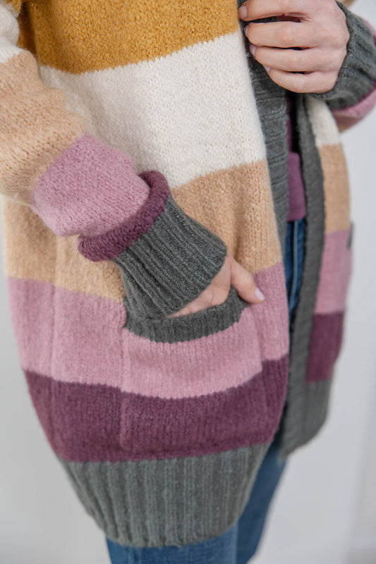 A close-up of a woman wearing a chunky knit cardigan with wide horizontal color-block stripes in mustard yellow, cream, beige, dusty pink, mauve, and gray. The cardigan has ribbed cuffs and a ribbed hem, and the person is also wearing blue jeans.