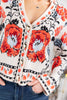 A close-up of a woman wearing a cream knit cardigan with large red and light blue floral motifs, black vine patterns, and decorative borders.