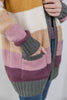 A close-up of a woman wearing a chunky knit cardigan with wide horizontal color-block stripes in mustard yellow, cream, beige, dusty pink, mauve, and gray. The cardigan has ribbed cuffs and a ribbed hem, and the person is also wearing blue jeans.