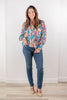 A woman stands in a bright room wearing a long-sleeved button-up blouse with an abstract paisley pattern in blue, orange, purple, and beige, blue jeans, and beige heels.