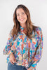A smiling woman with long brown hair wearing a colorful, long-sleeved button-up blouse with an abstract paisley pattern in blue, orange, purple, and beige, paired with blue jeans, against a plain white studio background.