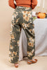 A woman shown from the back wearing wide-leg floral pants in olive green with large cream and tan flowers, paired with an orange ribbed sweater and brown sandals, standing indoors near a draped table with pumpkins and candles.