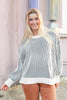 A smiling blonde woman wearing a Halloween inspired look with white knit sweater with thin black vertical stripes and orange pants, standing outdoors against a soft pink wall.