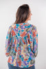 A woman viewed from the back wearing a colorful, long-sleeved blouse with a retro paisley pattern in blue, orange, purple, and beige, paired with blue jeans against a plain white background.