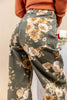 A close-up of a woman wearing wide-leg floral jeans in an olive green base with large cream and tan flowers, paired with an orange ribbed sweater.