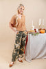 A woman models an outfit consisting of a tan short-sleeve V-neck top and dark olive green wide-leg pants with a beige and white floral pattern. She is standing in a studio setting next to a styled table with autumn decorations.
