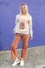 A woman stands in front of a purple brick wall wearing a cream-colored crewneck sweatshirt with a 'Pumpkin Season' graphic, paired with rust-colored cropped pants and white slip-on shoes.