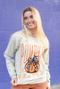 A smiling blonde woman wearing a cream-colored crewneck sweatshirt with a 'Pumpkin Season' graphic featuring an orange pumpkin and leopard print bow, posed against a purple painted brick wall.