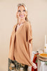 A smiling blonde woman models a loose, short-sleeve V-neck top in a warm camel tan color, paired with floral pants. The background is a soft beige studio setting with a styled table in the background holding a candle and a pie.