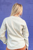 A woman with blonde hair is shown from behind wearing a cream-colored crewneck sweatshirt and brown pants, standing against a blue painted brick wall.