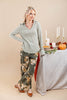 A woman models a light sage green ribbed knit long-sleeve top and matching wide-leg floral print pants with brown pointed-toe flats, standing next to a decorated table with candles and a pumpkin against a beige studio background.