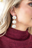A close-up of a woman wearing a triple-drop dangle earring with white circular charms featuring simple smiley faces, connected by gold-toned chains.