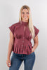 A woman modeling a cap-sleeve, high-neck peplum blouse in a dusty rose color, paired with blue jeans and brown ankle boots, standing against a plain white wall.