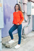 A woman posing outdoors in a bright orange Halloween crewneck sweatshirt, dark blue skinny jeans, and white sneakers, standing on concrete steps against a purple wall with industrial pipes.