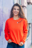 A woman smiling and looking to the side while wearing a bright orange crewneck sweatshirt with embroidered halloween graphics on the chest and sleeve, paired with blue jeans, set against a purple-toned urban wall background.