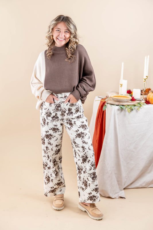 A woman modeling a brown and cream color blocked mock-neck knit sweater, paired with white wide-leg pants featuring a brown floral pattern, standing in front of a decorated table on a beige background.