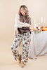 A woman modeling a cozy fall outfit consisting of a brown knit sweater, a cream knit cardigan, and white wide-leg pants with a dark floral pattern, standing in front of a warm beige background with a styled autumn table in the background.